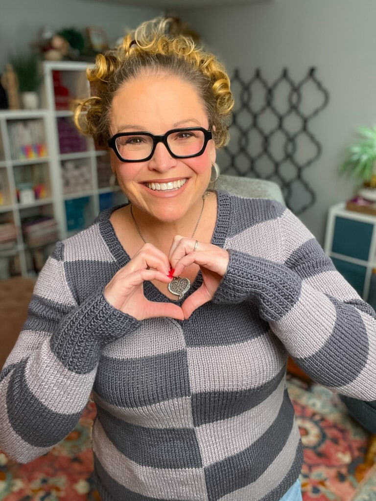 A smiling person with curly hair and glasses wears their favorite knit sweater pattern—a gray striped design—and forms a heart shape with their hands, holding a heart-shaped pendant, standing in a cozy, colorful room. -Marly Bird