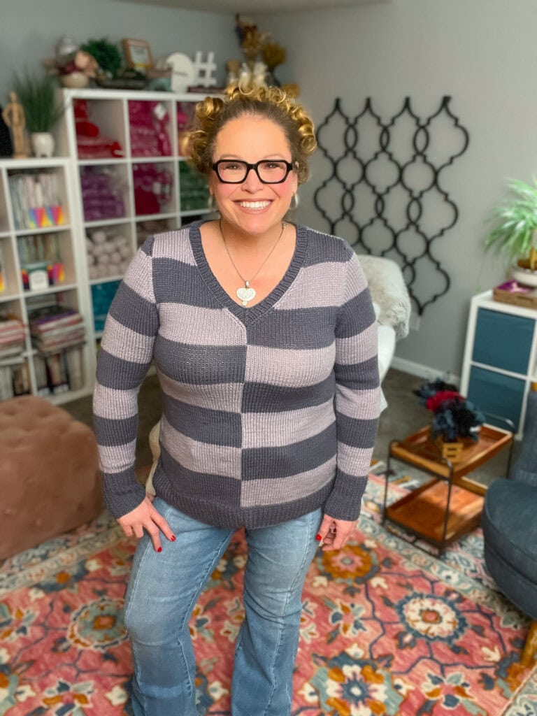A smiling woman with curly hair and glasses wears her favorite knit sweater pattern and jeans, standing in a colorful room with a patterned rug, bookshelves, plants, and decor. -Marly Bird
