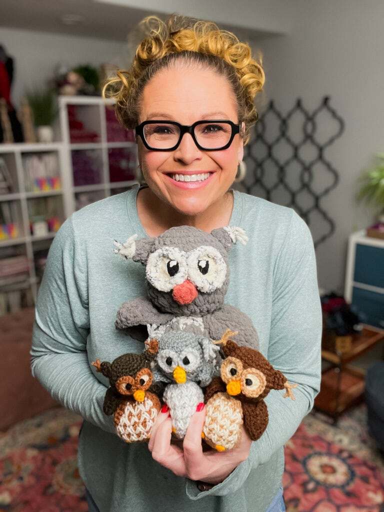 A smiling woman with curly hair and glasses holds several handmade crocheted owls in various colors, standing in a cozy room with shelves, plants, and free knitting and crochet patterns displayed in the background. -Marly Bird
