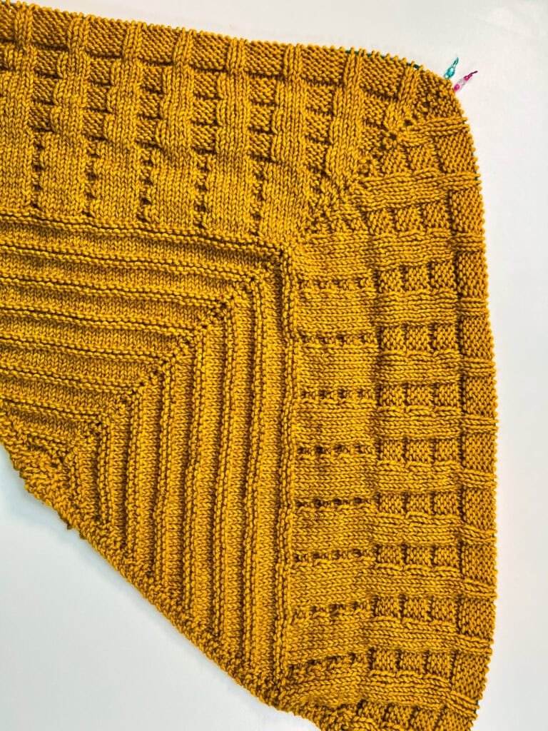 Mustard yellow knitted blanket featuring geometric textured stitches and a ribbed border, shown flat on a white background.