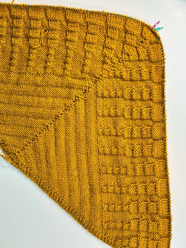 Mustard yellow knit fabric with square and stripe textures, triangular section, colorful stitch markers at top right on white background.