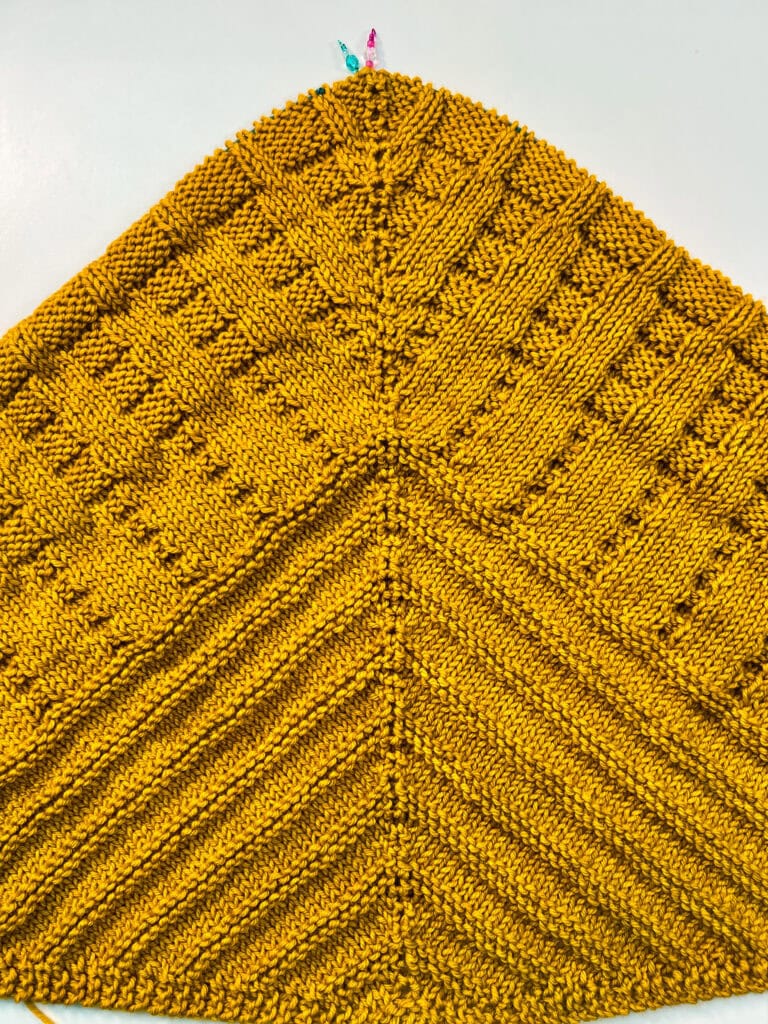 Yellow knitted triangular shawl with textured stitch patterns, symmetrical lines, and bright stitch markers attached.