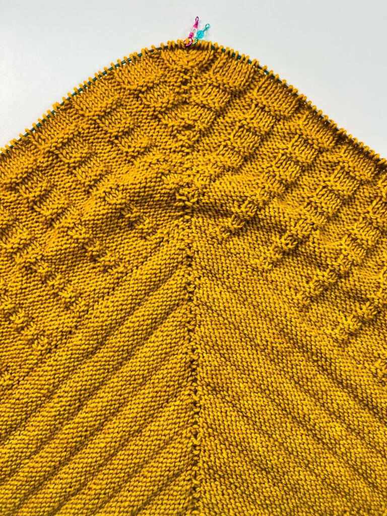 Yellow knitted panel with geometric textured patterns, central seam, and colorful stitch markers along the top edge.