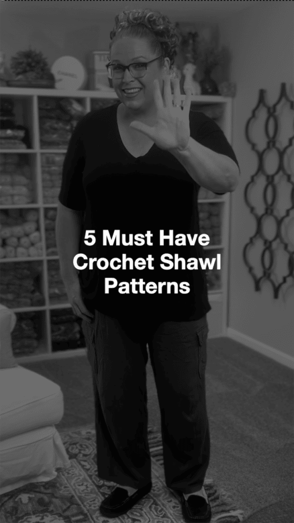 A smiling woman wearing glasses and a dark outfit stands indoors, waving. Shelves with yarn are behind her. The text on the image reads, "5 Must Have Crochet Shawl Patterns by Marly Bird. -Marly Bird