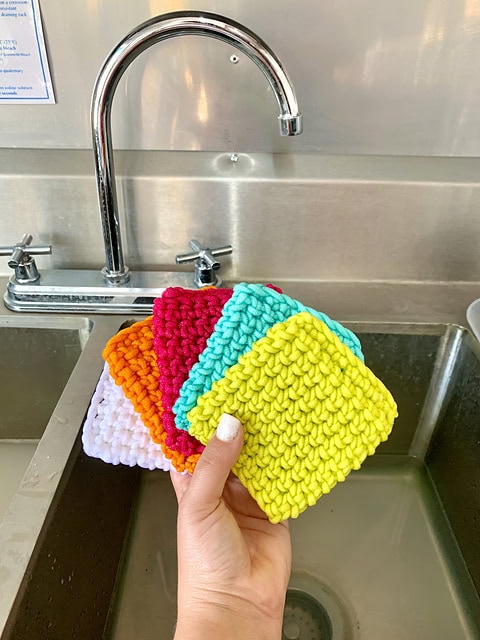 A hand showcases five vibrant crochet dishcloths—yellow, turquoise, red, orange, and white—in front of a stainless steel sink and faucet, perfect for spring cleaning tasks. -Marly Bird