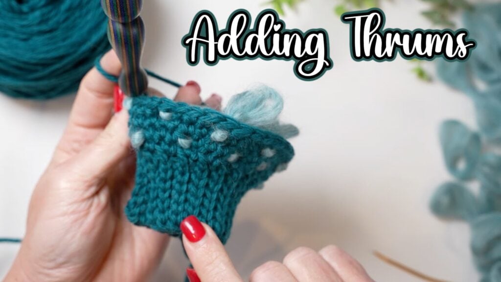Close-up of hands holding teal knitted fabric with added fluffy thrums, yarn ball in the background. Text overlay reads "Adding Thrums." Person has red nail polish—perfect inspiration for a fleece-lined crochet or Marly Bird’s crochet thrummed mitten pattern. -Marly Bird