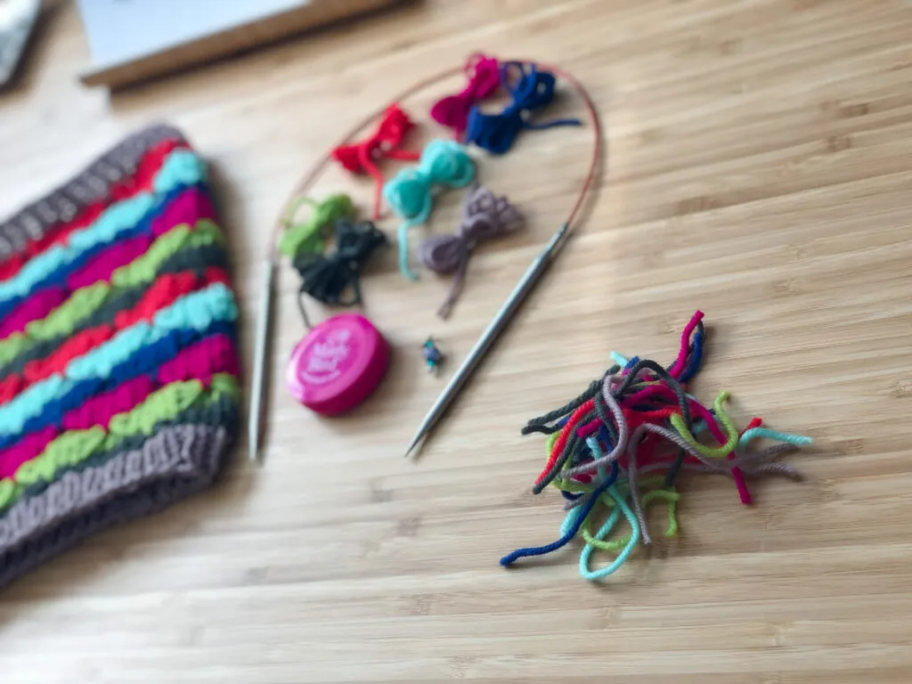Colorful yarn scraps and knitting materials, including circular needles, a striped knit project—perfect for a new Knit Hat Pattern—a pink tape measure, and Marly Bird stitch marker, are spread on a light wooden surface. -Marly Bird