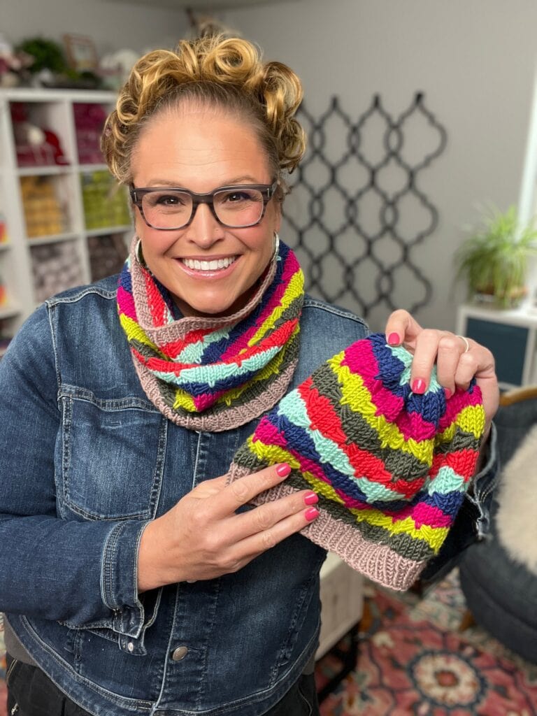 A smiling person wearing glasses, a denim jacket, and a colorful knit scarf holds up a matching Quickie Knit Hat in a cozy, well-lit room with shelves of yarn and craft supplies in the background. -Marly Bird