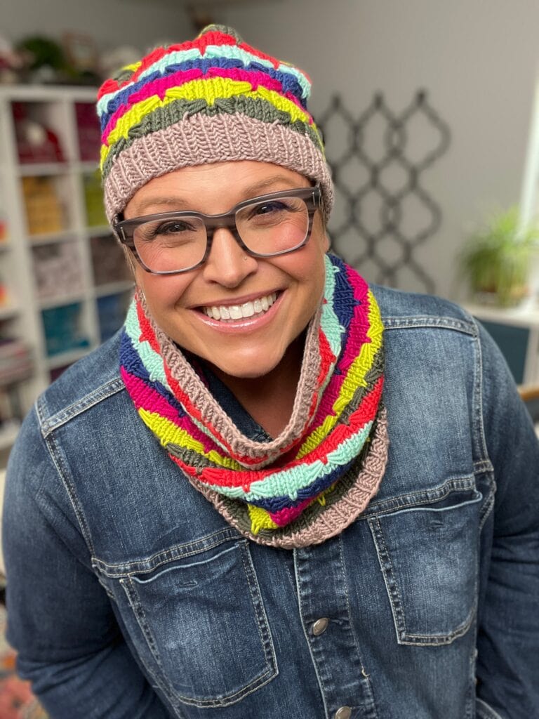 A smiling woman wearing glasses, a colorful knitted hat, and an Afternoon Delight Quickie Knit Cowl stands indoors in front of shelves filled with yarn. She is dressed in a denim jacket. -Marly Bird