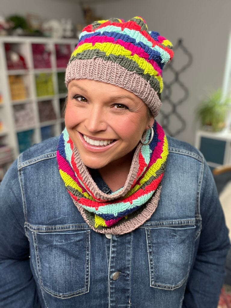 A smiling woman wearing a colorful Quickie Knit Hat and matching scarf stands indoors in front of shelves filled with yarn, dressed in a blue denim jacket. -Marly Bird