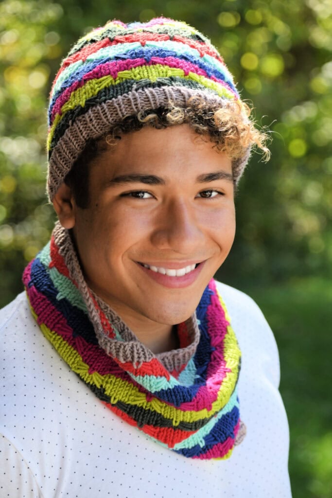 A young person with curly hair smiles outdoors, wearing a colorful Quickie Knit Hat and matching cowl scarf. Green trees and sunlight create a blurred background. -Marly Bird