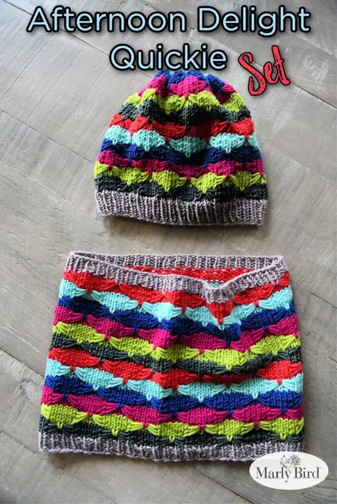 A colorful Quickie Knit Hat and cowl set with a bright geometric chevron pattern in blue, red, green, yellow, purple, and black, both with gray ribbed edges, displayed on a wooden surface. Text reads "Afternoon Delight Quickie Set. -Marly Bird