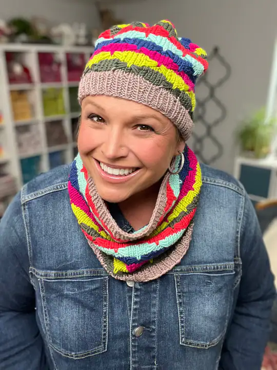 A smiling person wears a colorful knit hat and matching scarf with bright stripes over a denim jacket. Shelves with yarn are visible in the background, perfect inspiration for trying out new free knitting and crochet patterns. -Marly Bird