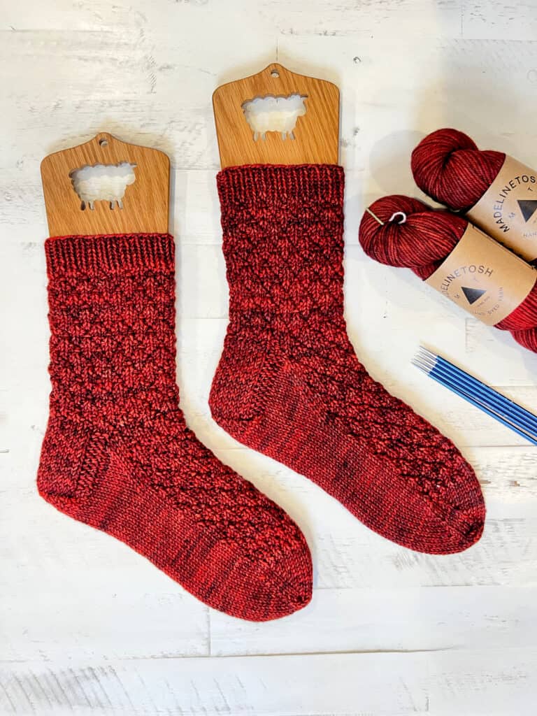 Two intricate toe-up knit socks are elegantly displayed on sock blockers. Beside them, matching red yarn balls and a set of knitting needles rest on a light wooden surface, inviting you to embark on sock knitting part 1 for your next cozy project. -Marly Bird