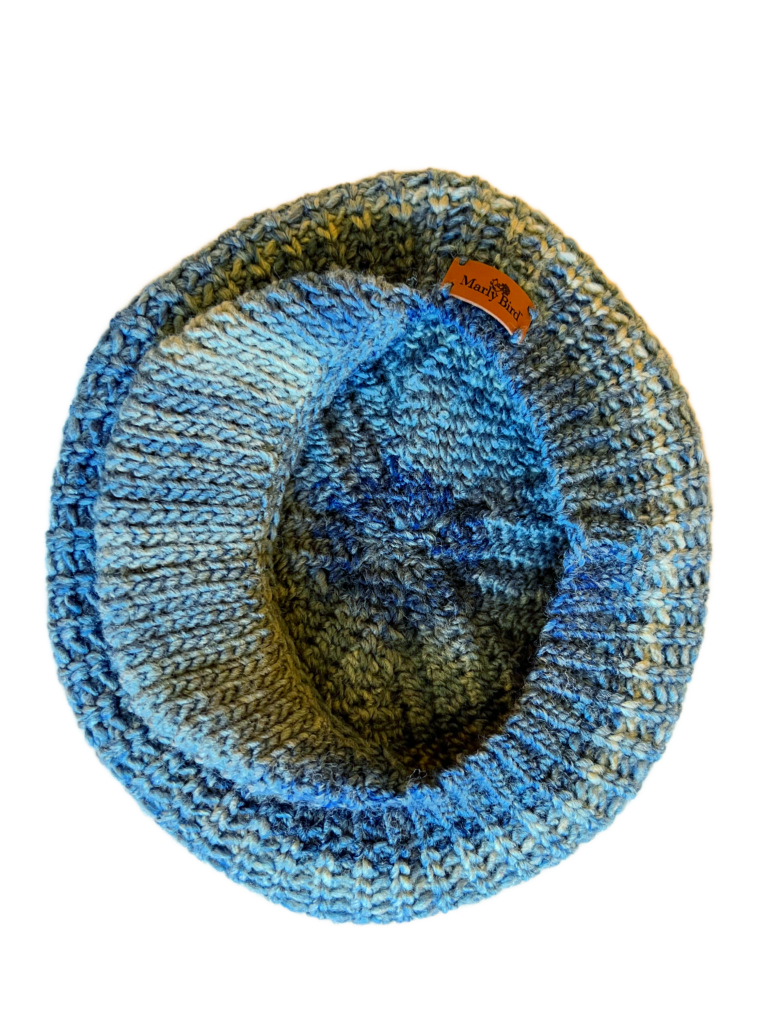 Inside look at this crochet hat featuring a gradient of blue hues, ranging from a soft sky blue to a deeper, more vibrant shade. The hat's intricate stitchwork is prominently displayed, showcasing the craftsmanship and skill involved in its creation. The interior of the hat reveals a rich texture, enhanced by the play of colors. A small, rectangular leather tag with the inscription "MarlyBird" is affixed to the hat, serving as a mark of its origin or brand. The black background contrasts sharply with the hat's colors, emphasizing its design and drawing attention to the detailed work. The hat's curvature and form are clearly visible, offering a unique perspective on its construction and design.
