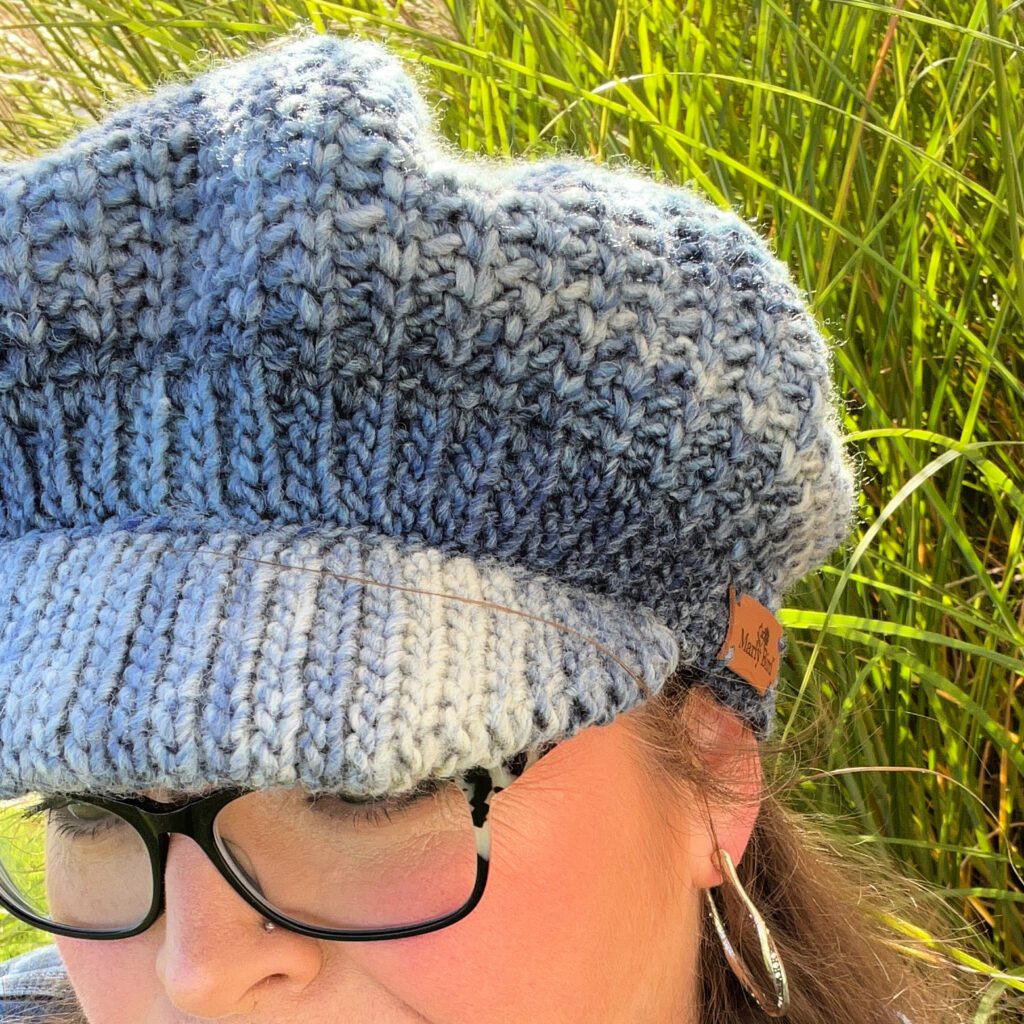 The image is a close-up view of a beautifully crafted crochet hat worn by a woman. The hat showcases intricate stitch detailing with an ombre effect transitioning from a lighter shade of blue to a darker one. The texture is rich and tactile, emphasizing the craftsmanship and attention to detail. Near the hat's brim, there's a small, rectangular leather tag with the logo or signature "Marly" engraved on it. The wearer's black-rimmed glasses are also visible in the shot, and a background of green, tall grasses adds a natural touch to the composition. The sunlight plays gently over the hat, highlighting its soft, cozy fibers and giving it a warm, inviting look. - marly bird
