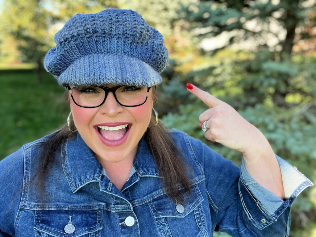Excited woman in denim jacket pointing to her blue crochet newsboy hat, surrounded by a green park setting. - marly bird