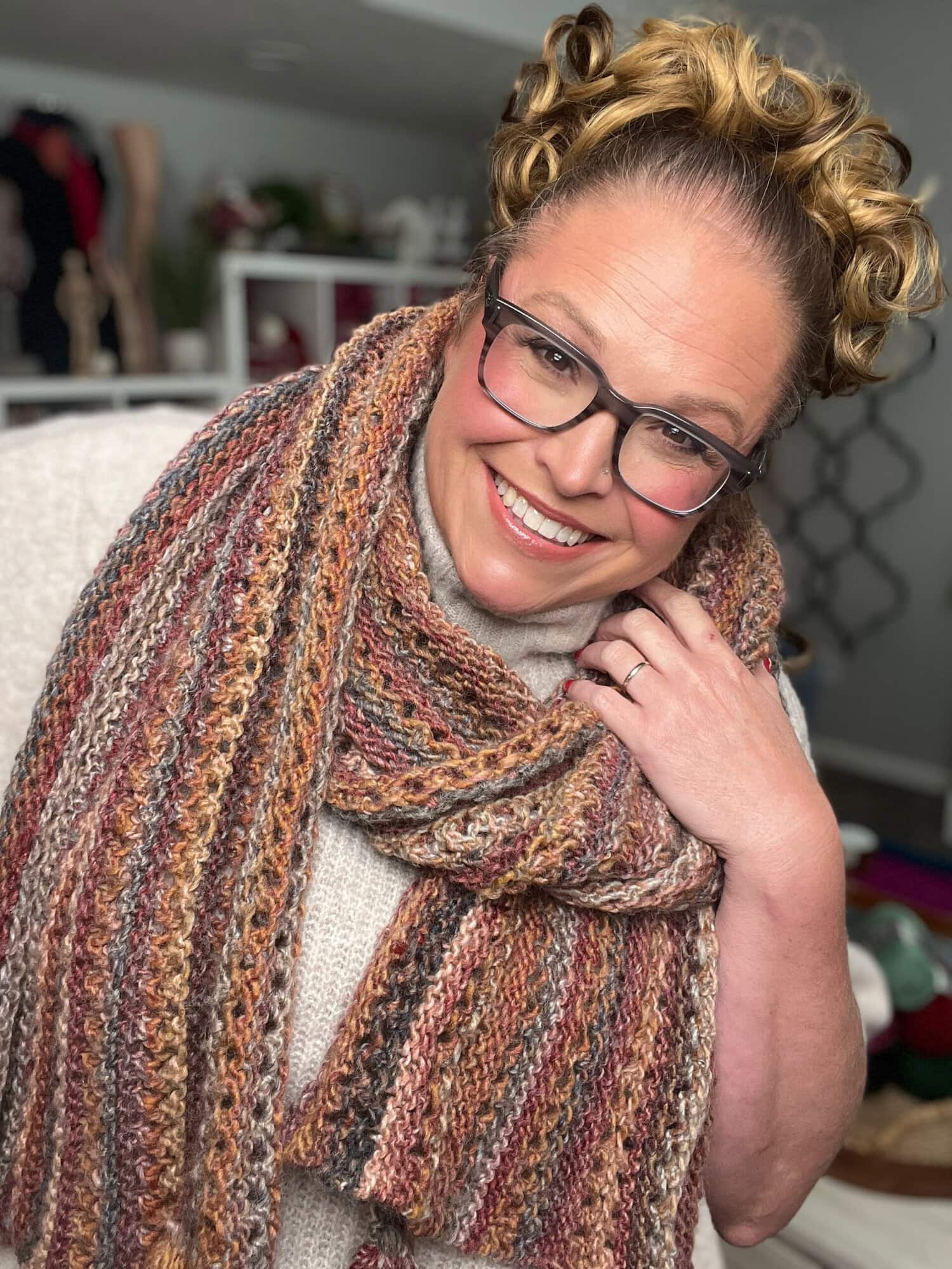 A woman models a cozy multicolored Almond Brittle knit wrap with visible Lace Bands stitch detail and corner tassels