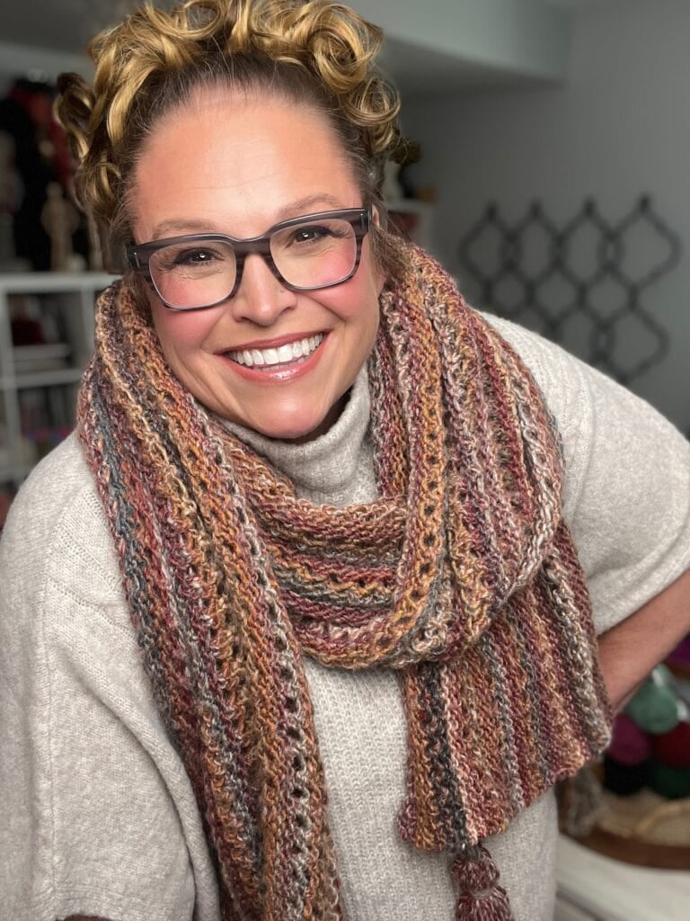 Person wears a handmade chunky knit scarf with multicolored yarn and tassels, showing varied stitches and cozy texture indoors.