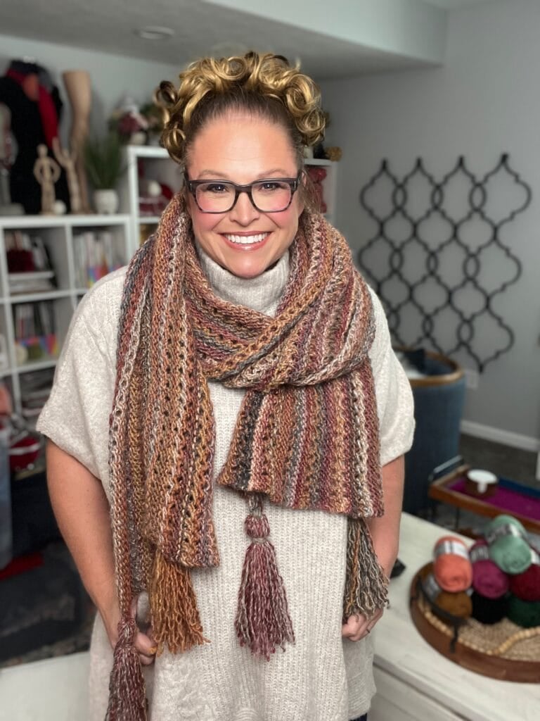 Marly Bird wearing the Almond Brittle free bulky knit lace wrap pattern in Caron Macchiato Cakes, with corner tassels and yarn supplies visible in a cozy crafting room background.