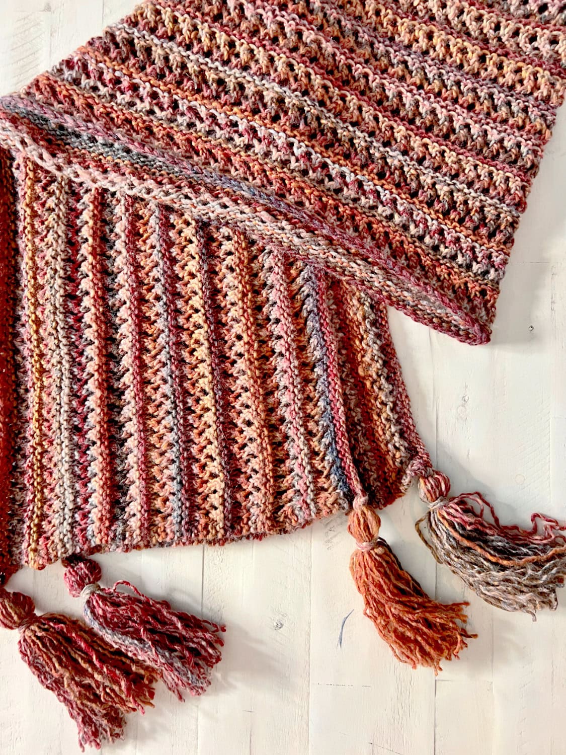 Close-up of the Almond Brittle Knit Wrap in Caron Macchiato Cakes, showing the textured striping in warm pink, peach, and brown colorway and the open Lace Bands stitch pattern