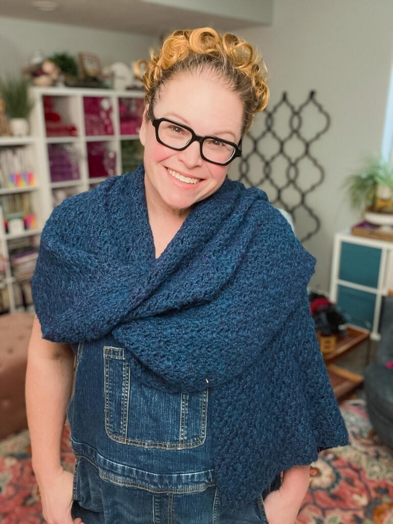 A woman models a textured blue alpaca crochet shawl, showing stitch detail; denim top and plants in the background.
