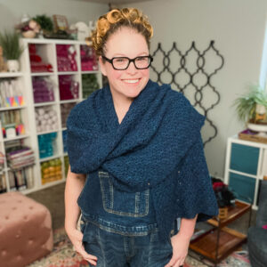 Textured navy blue crochet shawl draped over denim, showing stitch detail. Colorful yarn shelves and plants in the cozy background.