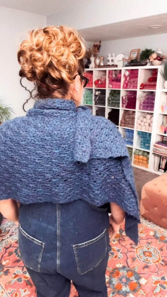 Curly-haired person wearing glasses models a blue crochet shawl, showing its textured stitches in front of shelves of colorful yarn.