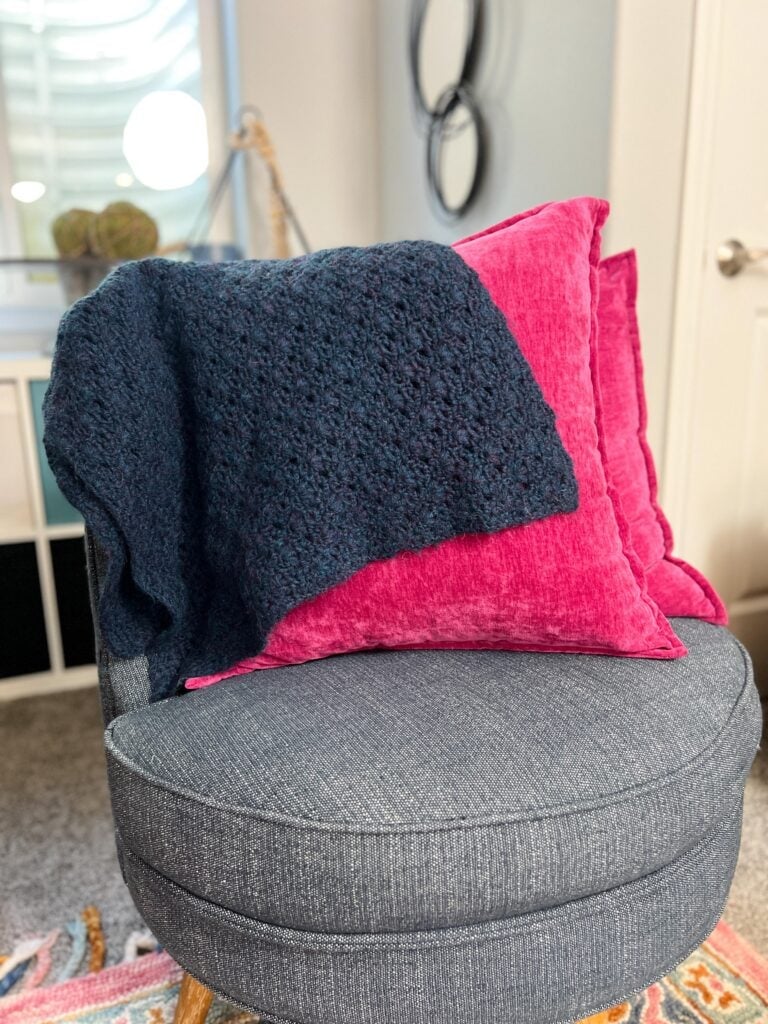Dark gray knitted blanket with visible stitch texture draped over pink pillows on a round gray chair in a bright, cozy room.
