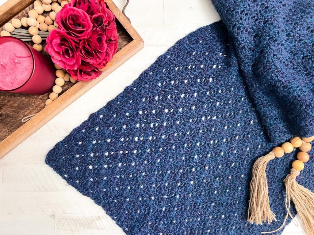 Deep blue crochet shawl with a textured pattern and tassels, displayed beside red roses and a candle on a wooden tray.
