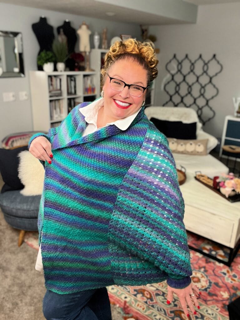 A woman with curly hair and glasses smiles while modeling Amanda Woods' colorful knit cocoon cardigan in shades of blue, purple, and green. She stands in a cozy living room with patterned rugs, bookshelves, and modern decor. Free knit pattern details are available nearby. -Marly Bird