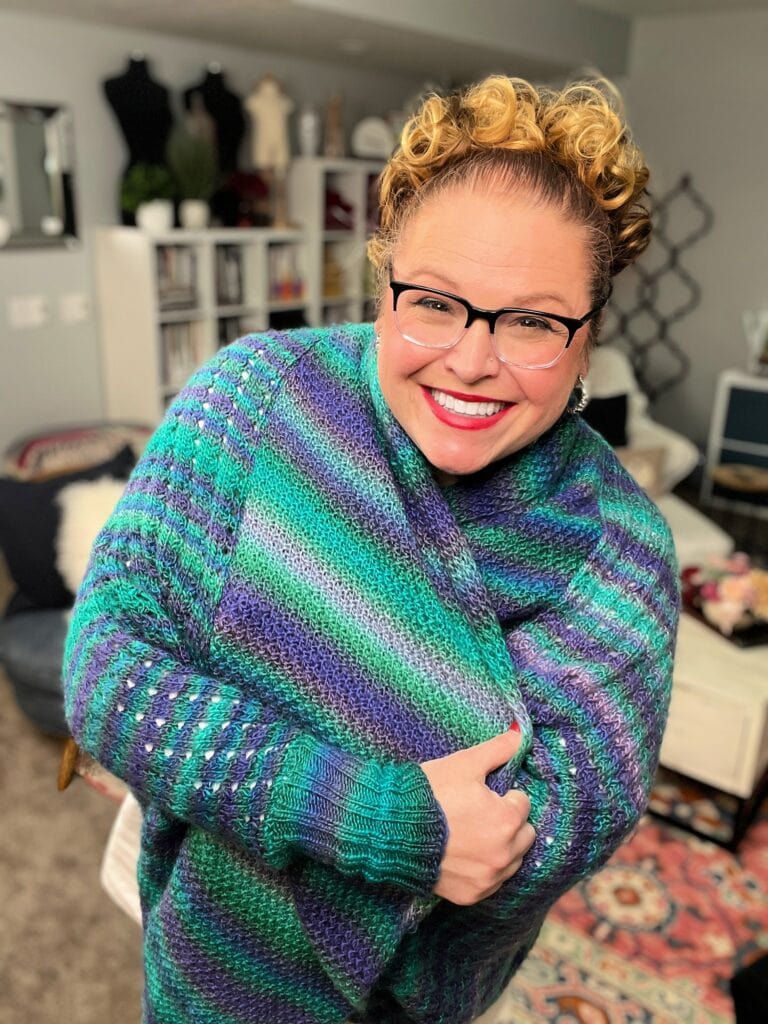 Amanda Woods, with curly hair and glasses, smiles brightly in a colorful blue and green striped knitted sweater. The cozy room's bookshelves add charm, just like her favorite knit cocoon cardigan pattern. -Marly Bird