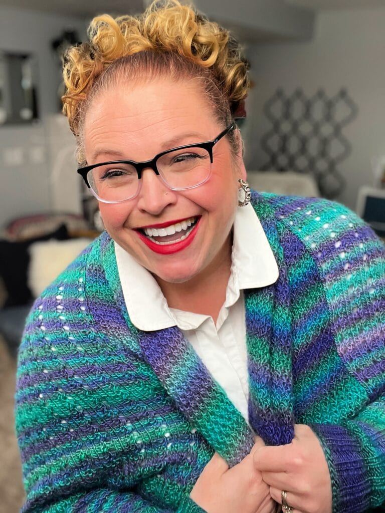 Amanda Woods, with curly hair and glasses, is smiling while wrapped in a vibrant knit cocoon cardigan. She is wearing a white collared shirt and standing in a cozy room with soft lighting. -Marly Bird