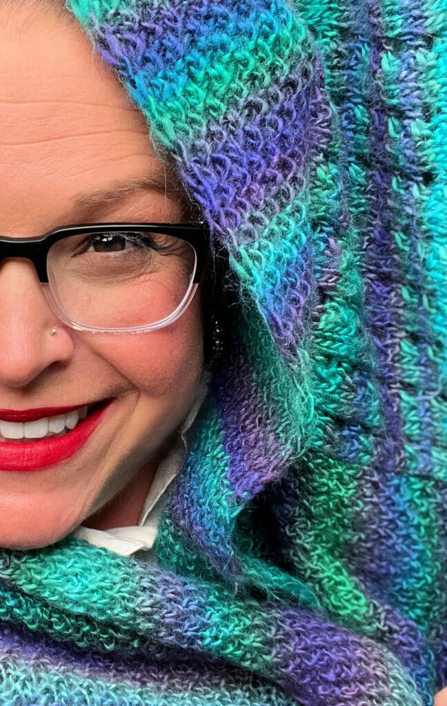 Close-up of a person smiling, wrapped in a vibrant green and purple knitted shawl that hints at Amanda Woods’ knit cocoon cardigan style. They have glasses and red lipstick, with part of their face covered by the cozy shawl. -Marly Bird