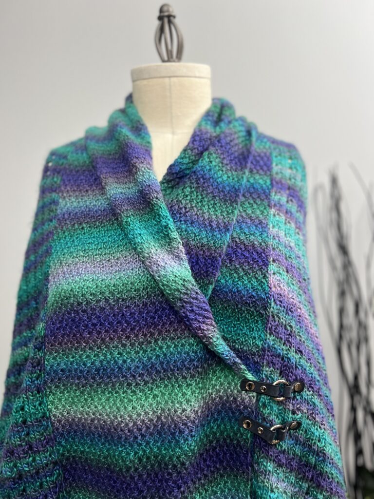 A mannequin showcases a multicolored shawl with shades of green, blue, purple, and gray. Fastened with a decorative clasp for a cozy, draped look reminiscent of the Amanda Woods style. The softly blurred background adds to its allure. -Marly Bird