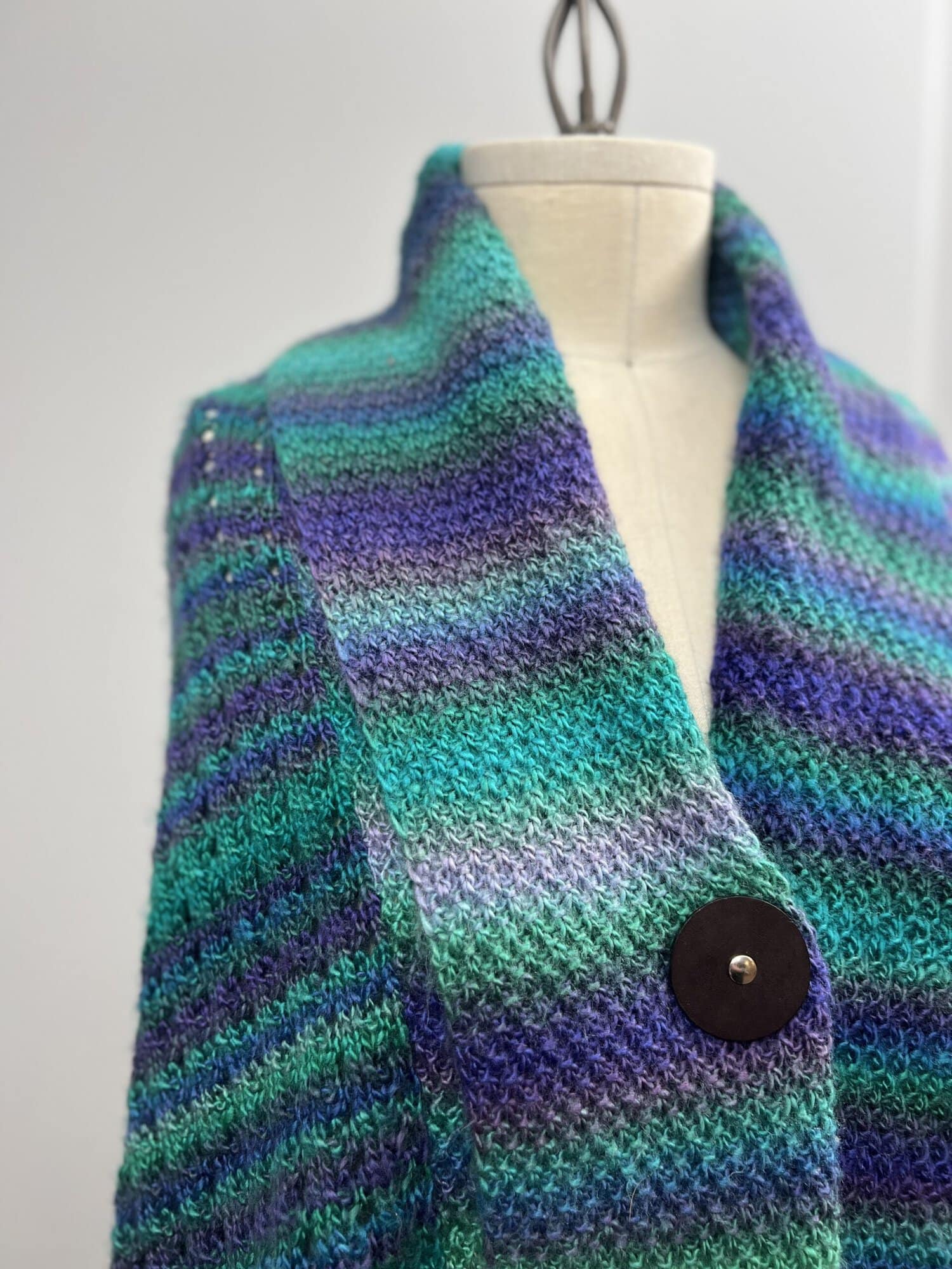 A close-up of a mannequin draped with a colorful, textured scarf featuring horizontal stripes in shades of blue, green, and purple. The scarf is fastened with a large, round button. It's reminiscent of Amanda Woods' Free Knit Pattern collection. -Marly Bird