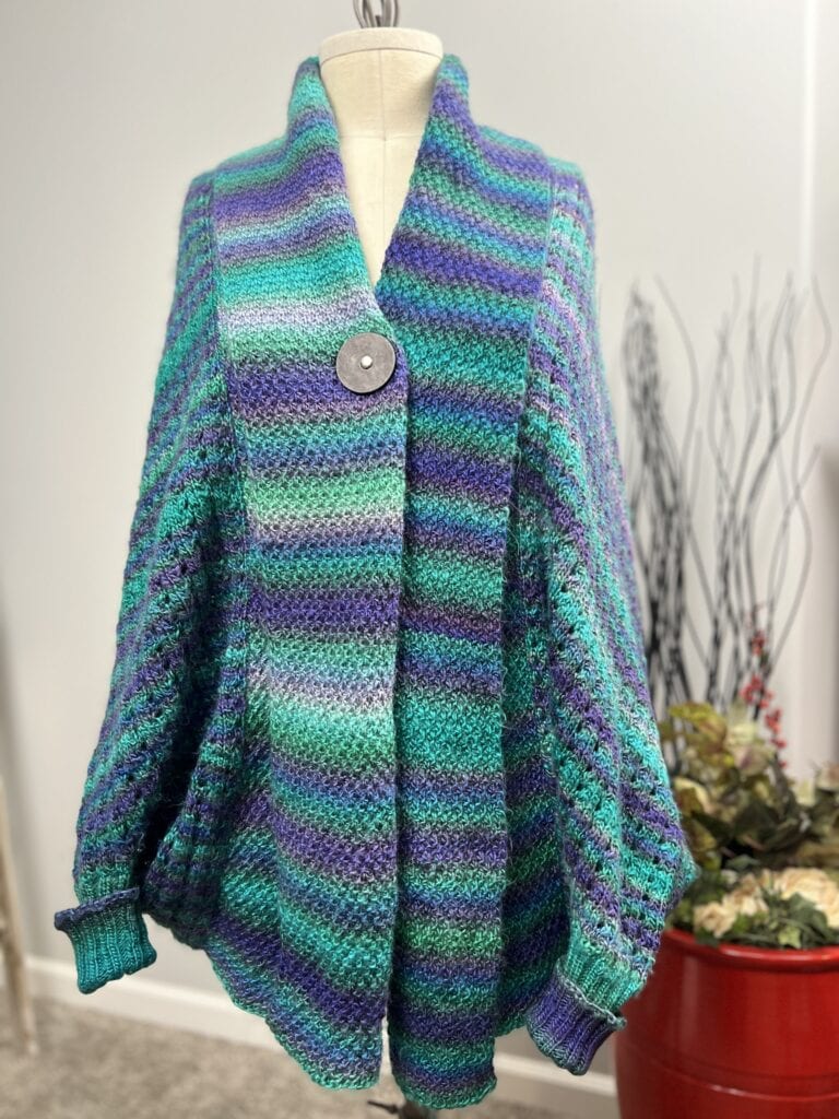 On display, the Knit Cocoon Cardigan—designed with a colorful blend of blue, purple, and green stripes—features a large button closure. The mannequin stands against a simple backdrop with a red vase and plants, offering inspiration for anyone seeking a free knit pattern. -Marly Bird