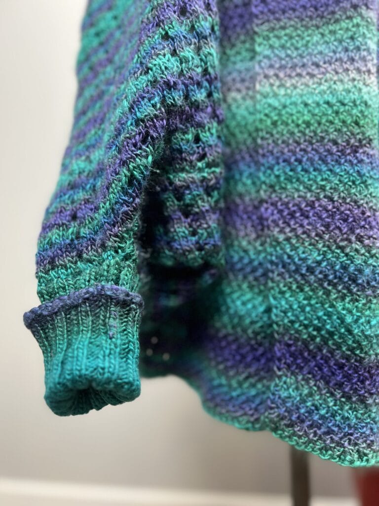 Close-up of a knitted sweater sleeve, featuring colorful horizontal stripes in shades of blue, green, and purple. Designed by Amanda Woods, the texture is woven with a ribbed cuff at the end. The background is softly blurred. -Marly Bird