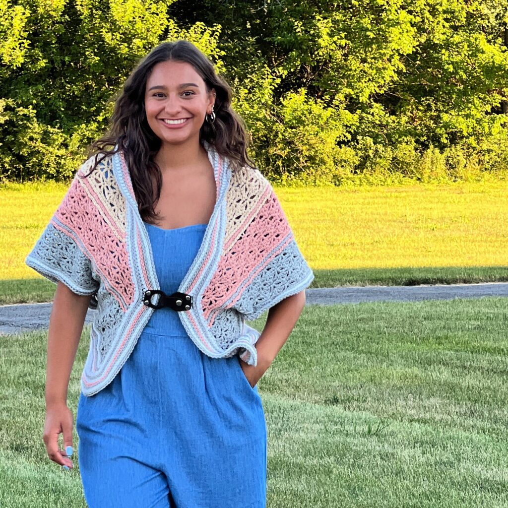 A woman smiles while walking on a grassy area in a blue dress and a stylish crochet shawl with pink, blue, and beige stripes fastened with a black clasp. Trees and a pathway are visible in the background. -Marly Bird