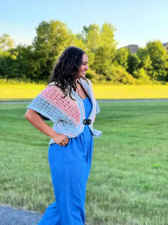 Woman standing on grass, wearing a blue jumpsuit and a pastel-colored cardigan made from free knitting and crochet patterns, smiling and looking to the side. Trees and a road are visible in the background on a sunny day. -Marly Bird