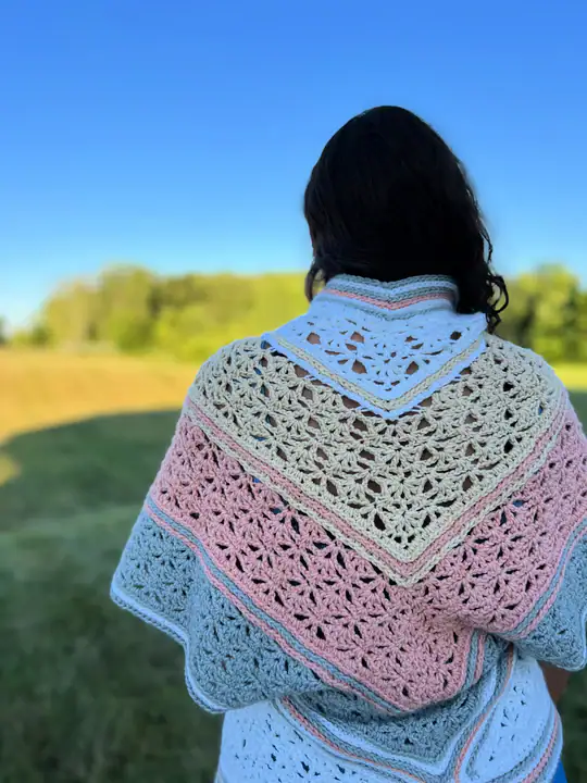 A person with long dark hair stands outdoors, facing away, wearing a crocheted shawl made from free knitting and crochet patterns in cream, pink, and blue tones. The clear blue sky and lush green trees complete the serene scene. -Marly Bird