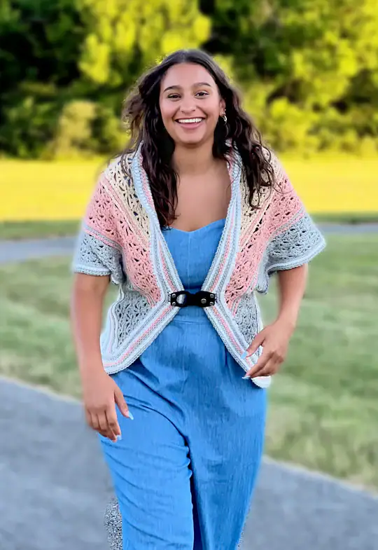 A smiling woman walks outdoors wearing a blue dress and a colorful knit shawl with pink, gray, and cream stripes—an inspiring look for those seeking free knitting and crochet patterns. Green grass and trees are blurred in the background. -Marly Bird