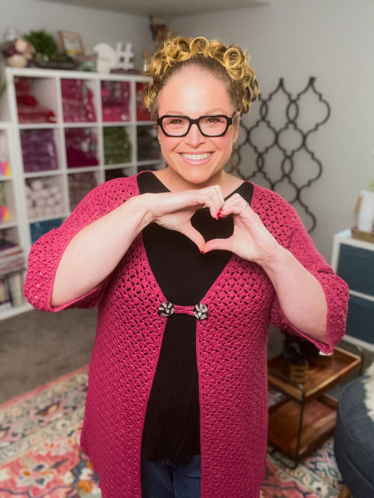 A smiling woman with glasses and curly blonde hair wears a pink Easy One Piece Crochet Cardigan, making a heart shape with her hands. She stands in a cozy room with shelves of yarn and home decor in the background. -Marly Bird