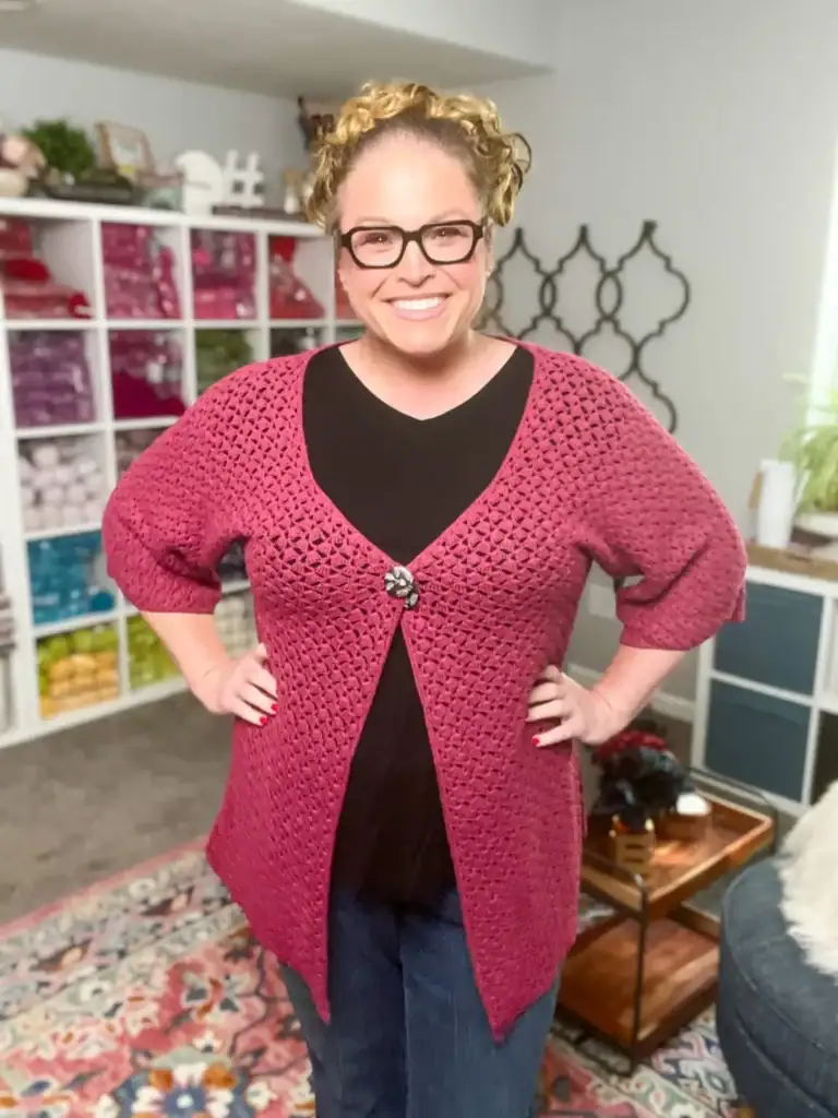 A smiling woman with curly hair in a bun, wearing glasses, a black top, and a pink crocheted cardigan stands in a cozy, well-lit room with shelves of yarn, free knitting and crochet patterns on display, and a patterned rug. -Marly Bird