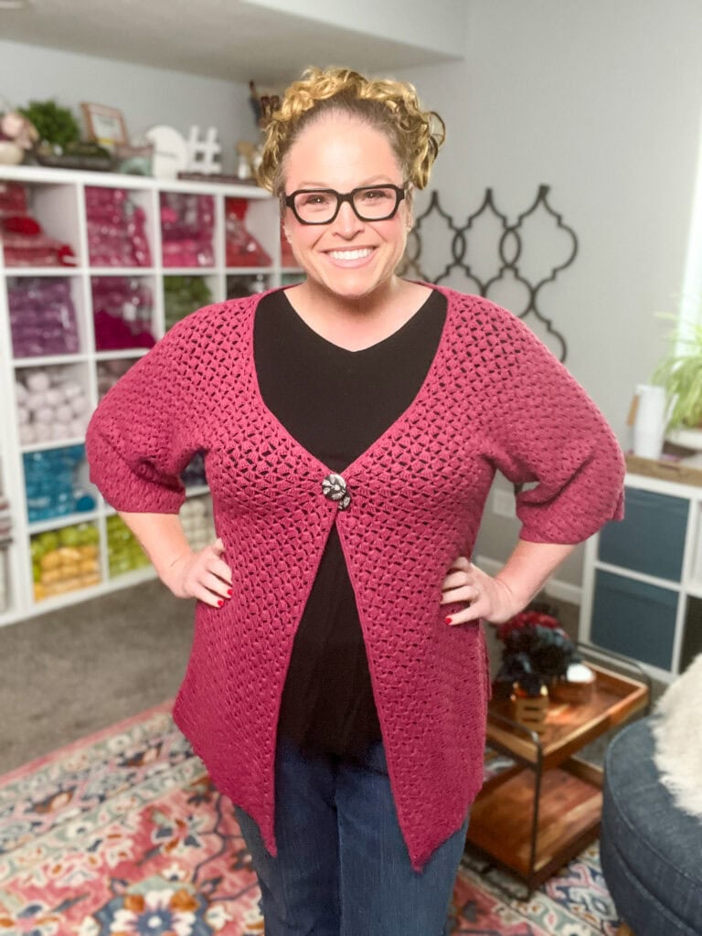 A smiling woman with curly hair wearing glasses and a deep pink crocheted cardigan, inspired by the Amimono Kimono Free Crochet Pattern, stands in a cozy room with colorful yarn shelves, a patterned rug, and home decor in the background. -Marly Bird