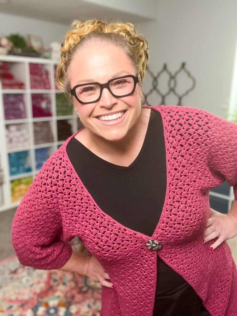 A smiling woman with blonde hair and glasses wears a pink crocheted cardigan—reminiscent of the Amimono Kimono Free Crochet Pattern—over a black top, standing in a colorful, yarn-filled room with hands on hips. -Marly Bird