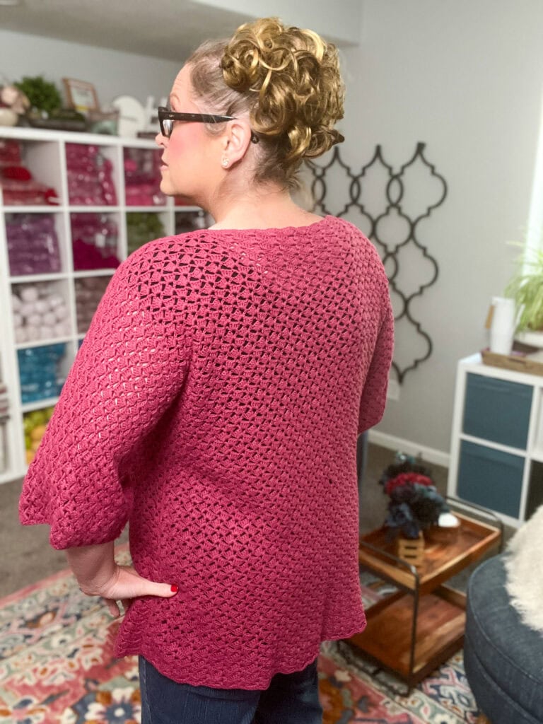 A woman with curly hair in an updo stands in a cozy, well-lit room, wearing a textured deep pink crochet sweater made with the Amimono Kimono Free Crochet Pattern. Shelves of yarn and a patterned rug are visible in the background. -Marly Bird
