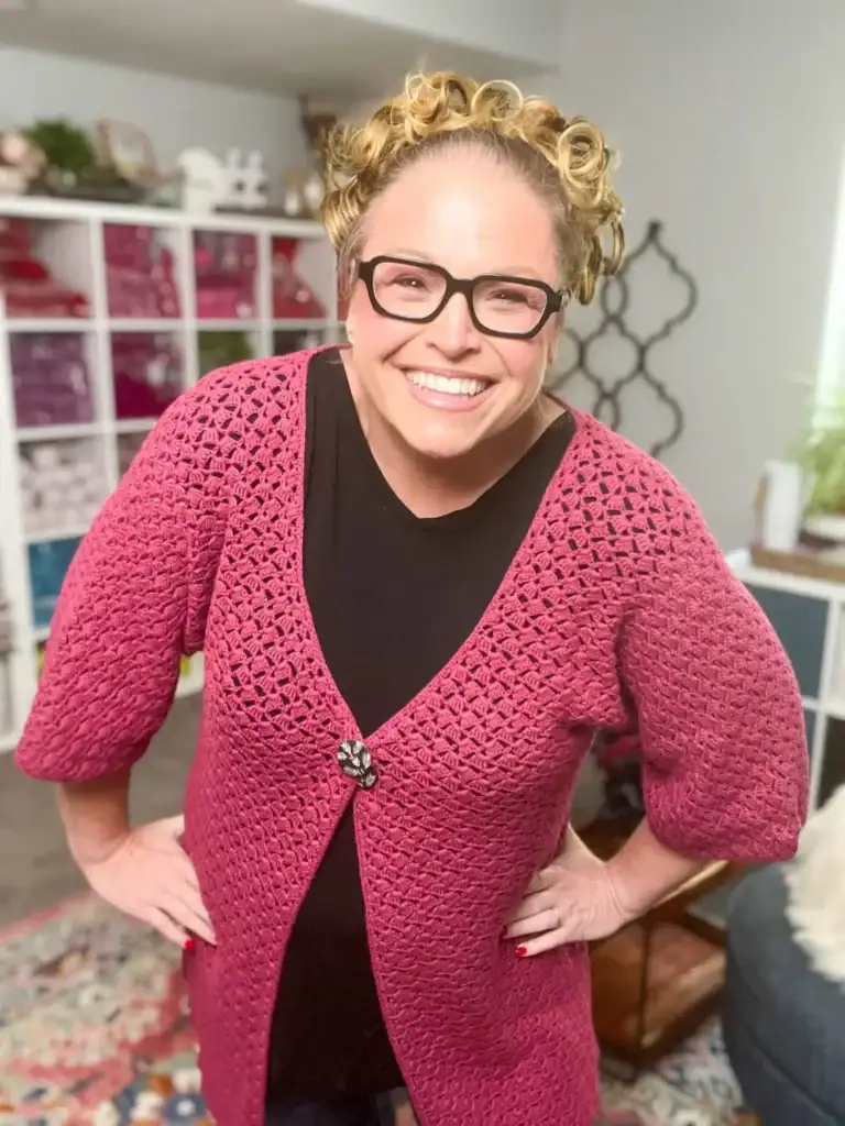 A smiling woman with light curly hair and glasses wears a pink crochet cardigan made from free knitting and crochet patterns over a black top, standing indoors with hands on her hips. Shelves and home decor are visible in the background. -Marly Bird