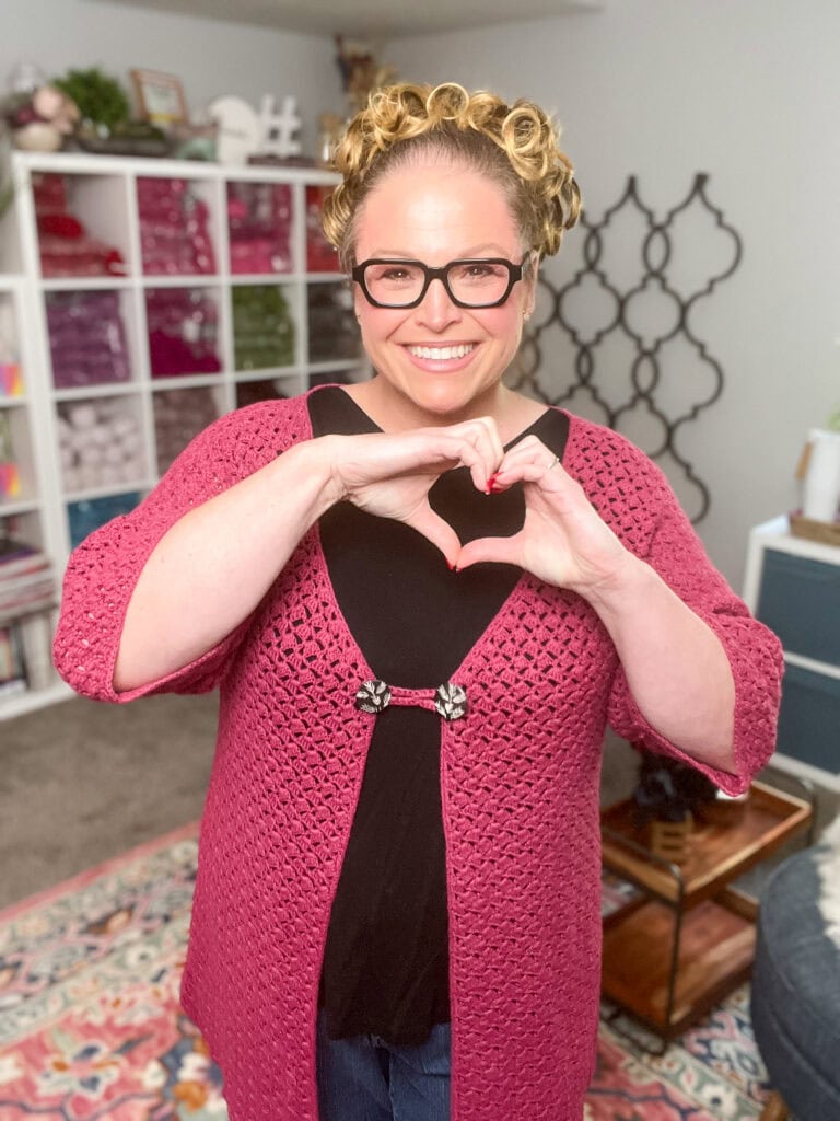 A smiling woman with curly blonde hair and glasses wears a pink crochet cardigan, inspired by the Amimono Kimono Free Crochet Pattern, over a black top. She stands indoors, forming a heart shape with her hands amid shelves and home decor. -Marly Bird