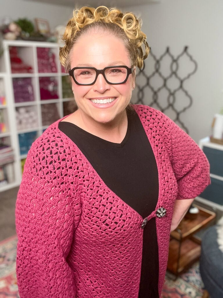 A smiling woman with glasses and curled hair wears a pink crocheted cardigan, reminiscent of the Amimono Kimono Free Crochet Pattern, over a black top. She stands in a cozy, well-lit room with shelves, books, and colorful decor in the background. -Marly Bird